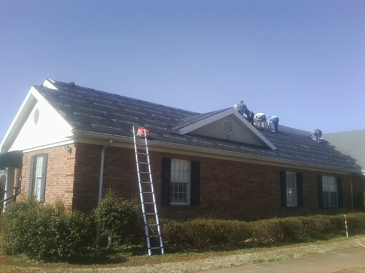 Crew tearing shingles off a brick ranch roof with a ladder set — Metal Roof Repair work by our crew in Francestown