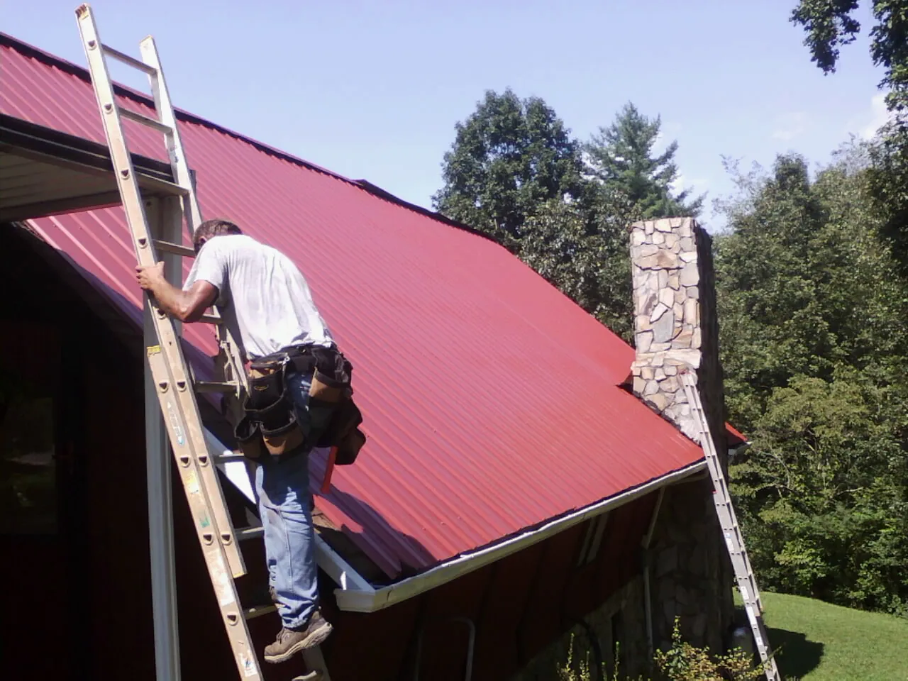 Roofer on a ladder beside a red metal roof and stone chimney — Soffit & Fascia Repair by Roofing Brothers in Francestown
